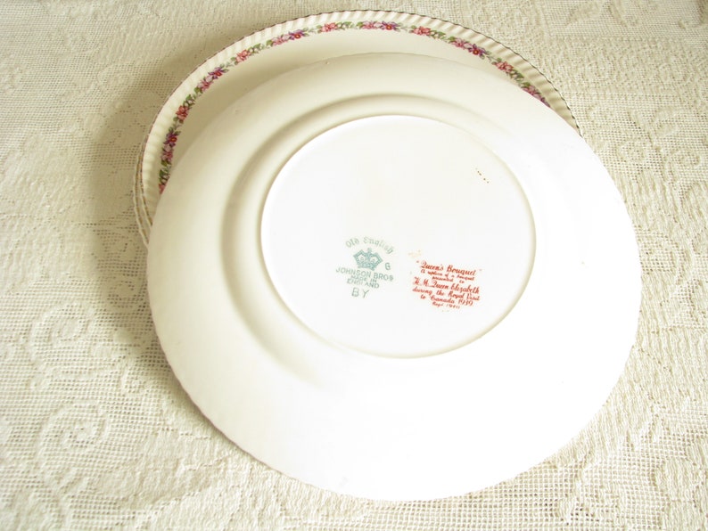 May include: A white ceramic plate with a floral border and a red and green inscription on the bottom. The inscription reads "Made in England, Johnson Bros. England,  'June Bouquet' a design by  J.H.  Town  Meredith during the Royal Visit to Canada, 1939."