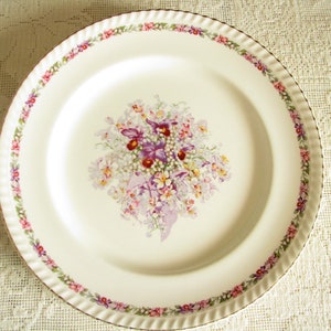 May include: A white ceramic dinner plate with a floral design. The plate has a scalloped edge and a gold rim. The floral design features purple, pink, and white flowers.