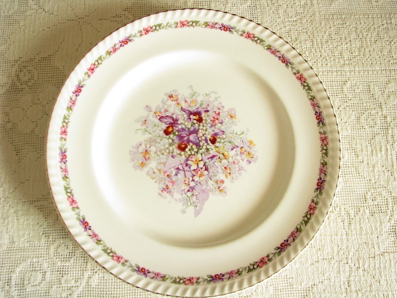 May include: A white ceramic dinner plate with a floral design. The plate has a scalloped edge and a gold rim. The floral design features purple, pink, and white flowers.