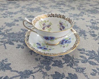 May include: A white teacup and saucer set with a floral design. The teacup and saucer have a gold trim and a pattern of blue flowers. The teacup has a curved handle. The saucer is round and has a matching floral pattern.