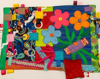 May include: A colorful baby sensory activity mat with various textures and elements, including flowers, a zipper, a key, a ring, and a small pillow. The mat is made of different fabrics and has a paisley pattern.