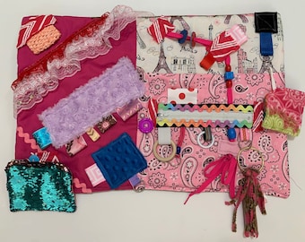 May include: A pink and white sensory activity mat with various textures and objects, including a zipper, a wooden ring, a fuzzy tassel, a sequin purse, and a button. The mat also features a pink and white paisley pattern, a purple fuzzy fabric, and a blue textured square.