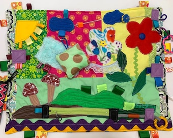 May include: A colorful baby activity play mat with various textures and shapes. It features a sun, mushrooms, a flower, a zipper, and a variety of ribbons and tags.