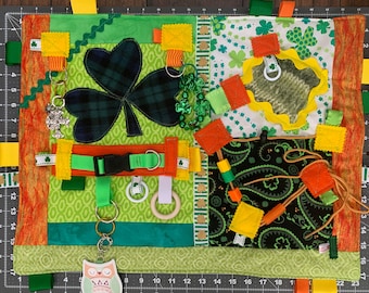 May include: A green, orange, and yellow baby activity mat with various textures and objects for sensory play. The mat features a large shamrock, a buckle, a wooden ring, a key chain with an owl charm, and a variety of ribbons and fabric squares.