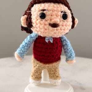 May include: A crocheted doll featuring brown hair, round wire-rimmed glasses, and black bead eyes. The figure wears a light blue long-sleeved shirt under a dark red vest, paired with tan trousers and white shoes, standing on a white circular base.