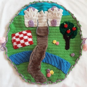 May include: A crocheted play mat with a castle, a tree with red apples, a picnic blanket, a brown path, and a blue river. The mat is surrounded by a green border with purple edging.