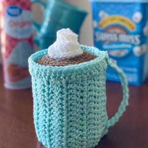 May include: A crocheted mug cozy in a light blue color, designed to resemble a cup of hot chocolate. The mug is topped with a brown crocheted layer and a white crocheted whipped cream. A can of whipped cream and a box of Swiss Miss are in the background.