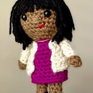 May include: A handmade crochet doll with black hair, brown skin, and a red smile. The doll wears a white jacket over a magenta dress. The doll's legs and arms are brown, and it has black shoes. The doll is a unique, handcrafted item.