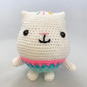 Op de afbeelding: Wit gebreid knuffeldier met zwarte ogen, een roze neus en een zwarte geborduurde mond. Het dier draagt een blauw en roze gebreide trui.