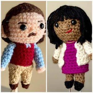 May include: Two handmade crochet dolls. One doll has a light skin tone, glasses, and a mustache, wearing a red vest, blue sleeves, and tan pants. The other doll has dark skin, black hair, and a white jacket over a magenta dress.
