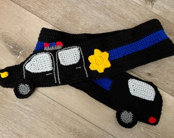 May include: Crocheted black and blue police car scarf with a yellow star. The scarf features a detailed police car design with a white roof and gray windows.