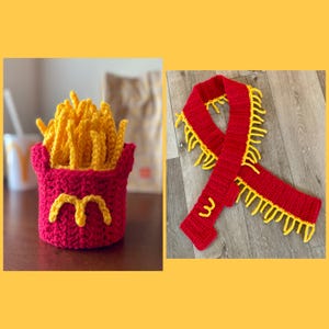 Op de afbeelding: Een gehaakte frietmuts in rood en geel. De muts is gevormd als een frietbakje en heeft een geel gehaakt frietontwerp bovenop. De muts ligt op een houten oppervlak.