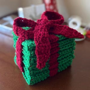 May include: A crocheted gift box with a red bow. The box is made of green and red yarn and is stacked in a square shape.