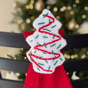 May include: A handmade Christmas tree-shaped kitchen towel holder. The white crocheted tree is decorated with red and green stitching. The holder is attached to a red towel with a textured pattern. The towel is hanging on a black chair.