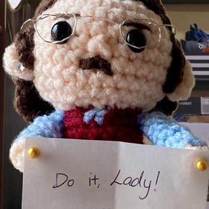 May include: A handmade crochet doll with a light skin tone, brown hair, and glasses. The doll wears a blue and maroon sweater and holds a note that says "Do it, Lady! ~Chit". The doll has black eyes and a small mustache.