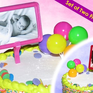 May include: A decorated cake with a pink frame holding a baby photo and a smaller cake with a family photo. Colorful round cake toppers in pink, green, yellow, and purple are also on the cake. Text reads "Set of Two Frames."