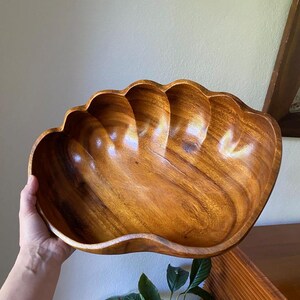 May include: A large, polished wooden bowl carved into the shape of a seashell. The bowl features a warm, medium-brown wood grain with visible natural patterns and a smooth, curved rim that mimics the scalloped edges of a clam shell.