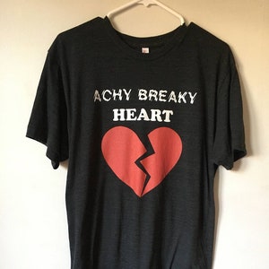 Puede incluir: Una camiseta negra con el texto "Achy Breaky Heart" y un gráfico de corazón roto rojo.