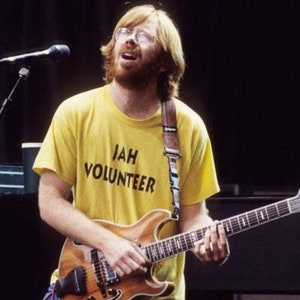 May include: A man with a red beard and glasses is playing a guitar. He is wearing a yellow t-shirt with the words "JAH VOLUNTEER" printed on it.
