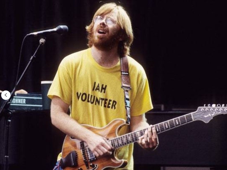 May include: A man with a red beard and glasses is playing a guitar. He is wearing a yellow t-shirt with the words "JAH VOLUNTEER" printed on it.