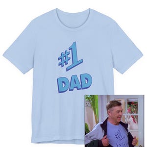 May include: A light blue t-shirt with the text "#1 DAD" in a blue, outlined font. The shirt has short sleeves and a crew neck. The image also includes a man wearing the shirt.