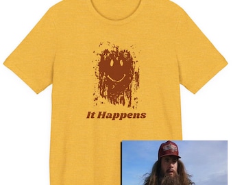Puede incluir: Camiseta amarilla mostaza con un diseño de cara sonriente marrón y el texto "It Happens". Una pequeña imagen de un hombre con pelo largo y barba está en la esquina inferior derecha, con el texto "IT HAPPENS."