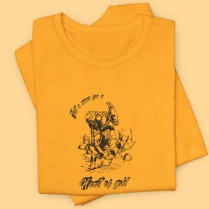Könnte beinhalten: Ein gelbes T-Shirt mit einer schwarzen Illustration einer Person, die mit einem Esel nach Gold sucht. Der Text "Just a miner for a Heart of gold" ist auf das T-Shirt gedruckt.