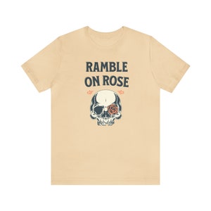 May include: A beige t-shirt with the text "Ramble On Rose" in black lettering. A skull with a rose in its eye socket is printed below the text.