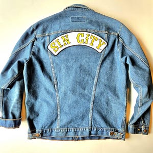 Op de afbeelding: Een lichtblauwe spijkerjas met een "SIN CITY" patch op de rug. De patch heeft gele letters op een witte achtergrond. De jas heeft een klassiek ontwerp met knopen en zakken.