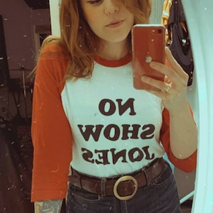 May include: A white and red baseball tee shirt with the text "JONES SHOW NO" printed in black. The person is wearing a brown leather belt with a large silver buckle.