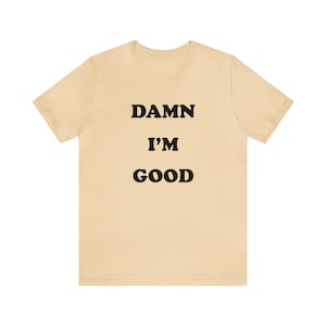 May include: A beige t-shirt with the text "DAMN I'M GOOD" printed in black.