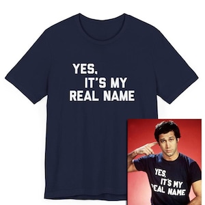 May include: A navy blue t-shirt with white text that reads "YES. IT'S MY REAL NAME". The shirt is shown on a model wearing the same shirt, with a red background.