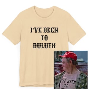 以下が含まれることがあります： ベージュのTシャツに「I've Been To Duluth」という黒い文字がプリントされています。テキストは太字のウエスタンスタイルのフォントです。右下には、同じようなシャツを着た男性の小さな画像が表示されています。