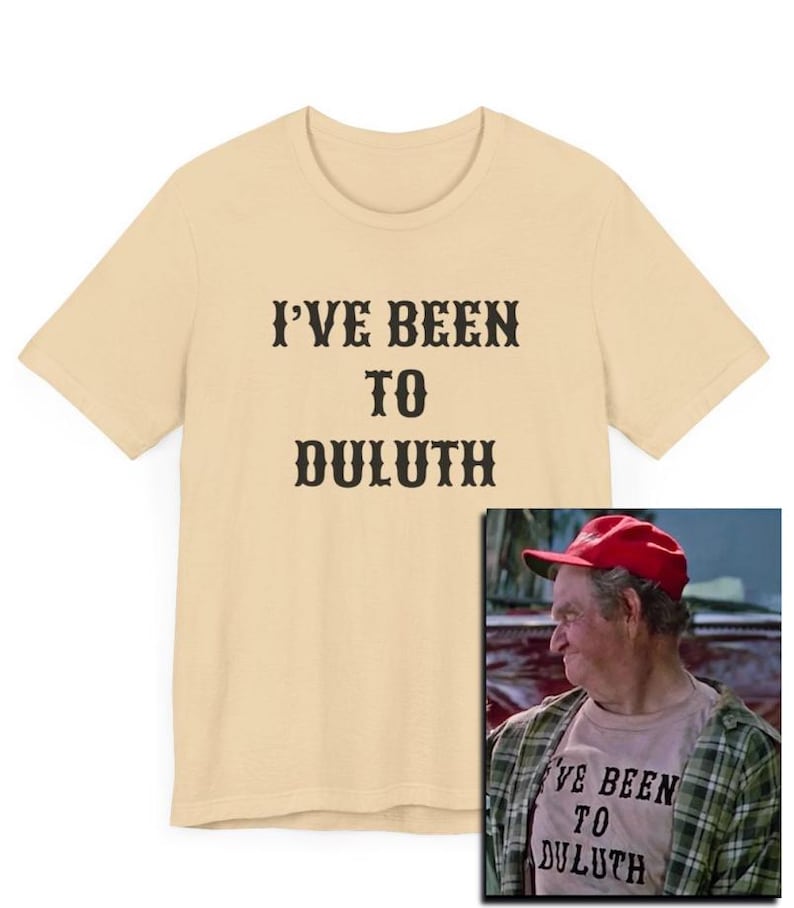 以下が含まれることがあります： ベージュのTシャツに「I've Been To Duluth」という黒い文字がプリントされています。テキストは太字のウエスタンスタイルのフォントです。右下には、同じようなシャツを着た男性の小さな画像が表示されています。