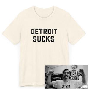 May include: Cream-colored t-shirt with the text "DETROIT SUCKS" in bold, black capital letters. The image also includes a black and white photo of a person flexing, wearing the same shirt.