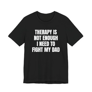 May include: Black t-shirt with white text that reads: "THERAPY IS NOT ENOUGH I NEED TO FIGHT MY DAD". The shirt has short sleeves and a crew neck.