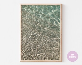 May include: A framed print featuring a photograph of clear, turquoise water with light reflections. The water's surface has a rippled texture, and the image is framed in a light-colored wooden frame. The print has a soft, natural aesthetic.