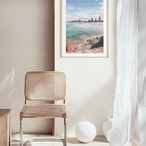 May include: A framed photograph of a beach scene with turquoise water, white sand, and a line of trees in the distance. The photo is hanging on a white wall in a room with a wicker chair and a white lamp.