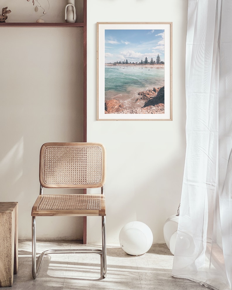 May include: A framed photograph of a beach scene with turquoise water, white sand, and a line of trees in the distance. The photo is hanging on a white wall in a room with a wicker chair and a white lamp.