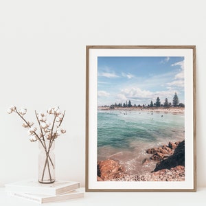 May include: A framed photograph of a beach scene with turquoise water, sandy shore, and a line of trees in the distance. The photo is in a light wood frame.