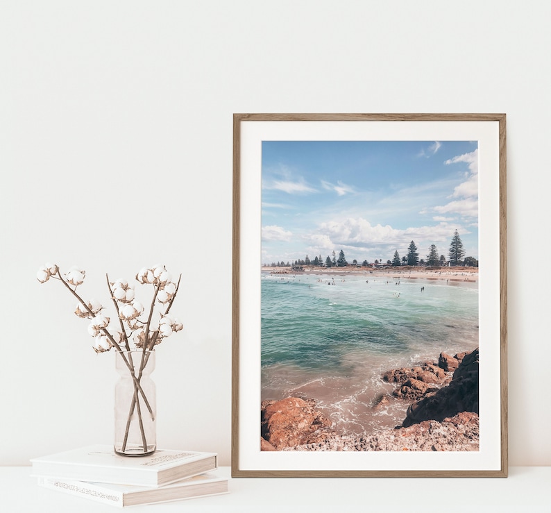May include: A framed photograph of a beach scene with turquoise water, sandy shore, and a line of trees in the distance. The photo is in a light wood frame.