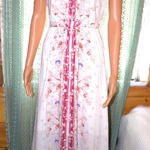 Peut inclure: Robe blanche &agrave; col licou avec un motif floral dans les tons de rouge, marron et bleu. La robe pr&eacute;sente une bande verticale de tissu magenta au centre et une taille cintr&eacute;e. La robe est longue.