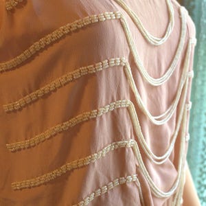 May include: A light pink dress with horizontal rows of white beads and cascading beaded strands. The dress has a scoop neckline and a soft, flowing fabric, creating a vintage or art deco aesthetic.