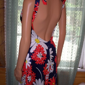 Könnte beinhalten: Ein bodenlanges Vintage-Kleid mit tiefem Rückenausschnitt und Bindeträgern. Das Kleid hat einen marineblauen Hintergrund mit einem Blumenmuster aus roten und weißen Gänseblümchen. Die Träger sind rot mit weißen Blumenakzenten.