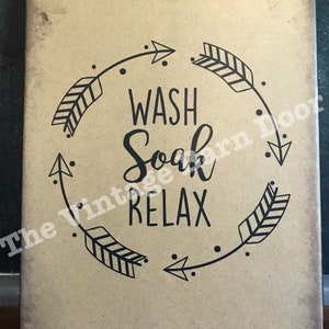 Puede incluir: Un letrero rectangular de madera color beige con texto negro y detalles de flechas. Las palabras "WASH, SOAK, RELAX" están centradas, rodeadas por un diseño circular de flechas. El letrero tiene una estética rústica y vintage.