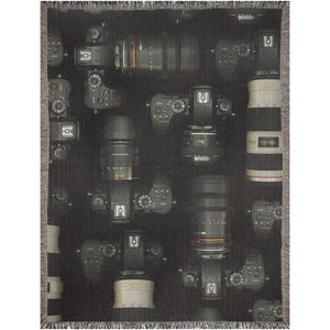 May include: A black and gray woven throw blanket featuring a repeating pattern of cameras and lenses. The design includes various camera models and lenses, creating a photographic theme. The blanket has a fringed edge.