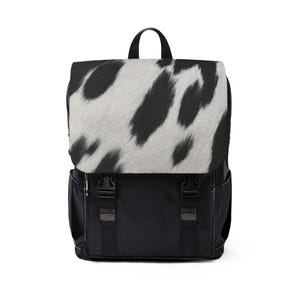 May include: A black backpack featuring a top flap with a white and black cow print pattern. The bag includes a top handle, two black side pockets, and two black adjustable straps with plastic buckle closures on the front main compartment.