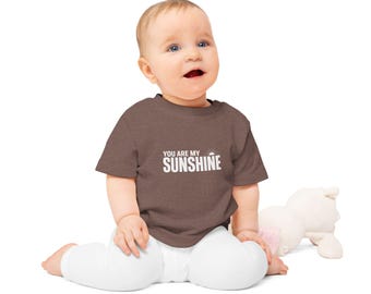 Może przedstawiać: Brązowa koszulka dla niemowlęcia z białym napisem "You are my sunshine" i małym graficznym słońcem. Niemowlę nosi koszulkę i siedzi na białym tle.