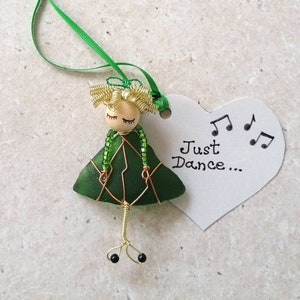 May include: A green glass figurine of a girl with blonde hair and closed eyes. She is wearing a green dress and has copper wire legs. The figurine is attached to a white heart-shaped tag with the text "Just Dance..." and musical notes.