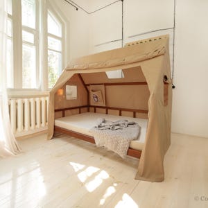 May include: A tan canvas tent-like bed frame with a wooden base and a white mattress. The bed is set up in a room with white walls and a white curtain.